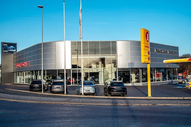 Moderne Porsche-forhandler med sølvfarget fasade og store glassvinduer, sett fra rundkjøring i dagslys.