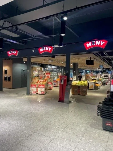 Inngang til MENY med tydelig skilt, handlekurver og frukt- og grøntavdeling innenfor.