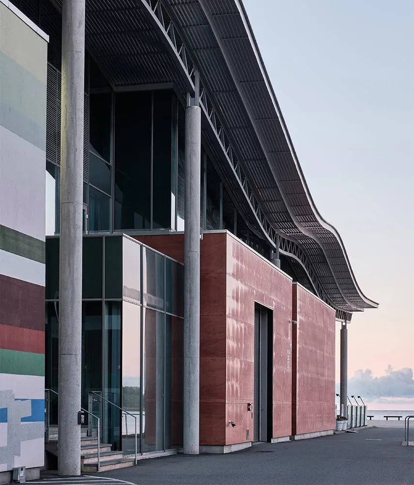 Bygning med bølgende tak, røde veggpaneler og store glassflater ved vannkanten i dempet kveldlys.