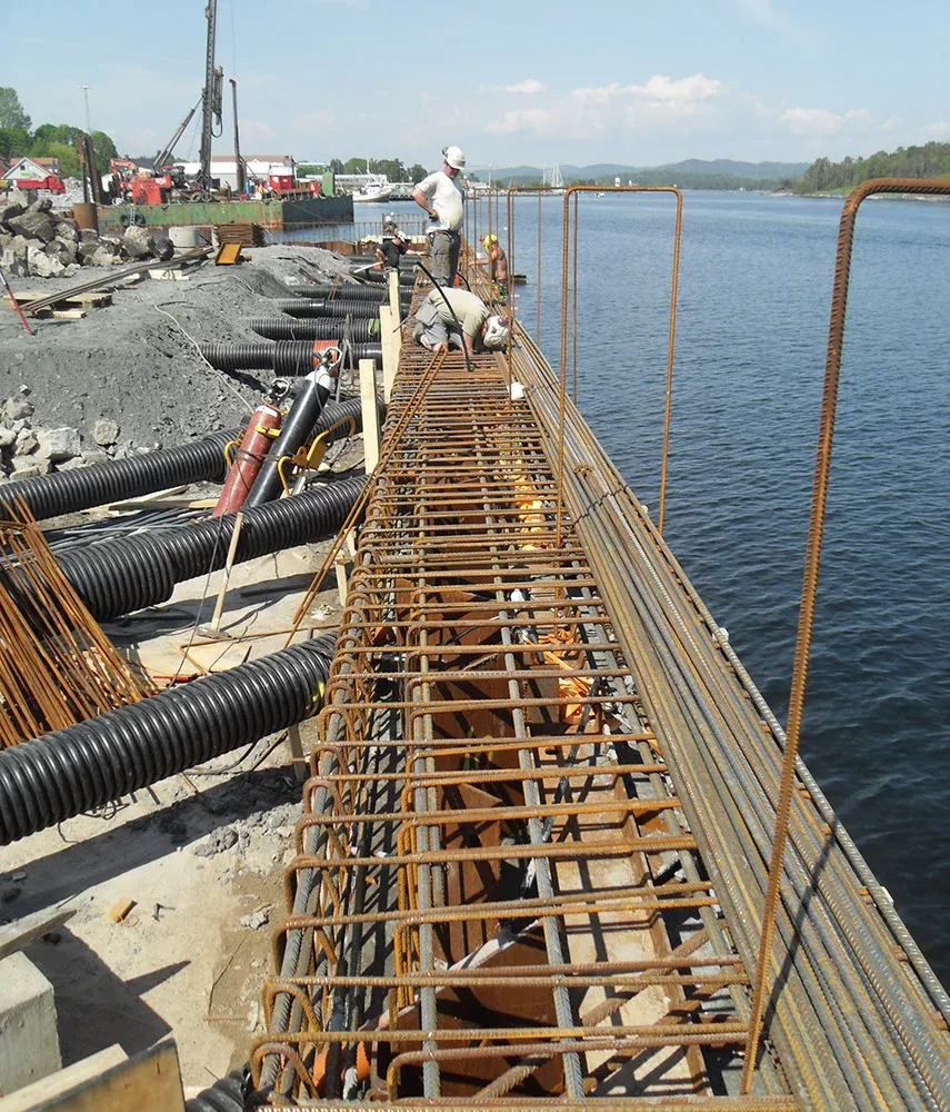 Arbeidere monterer armeringsjern og rør ved vannkanten under bygging av kai- eller bruinstallasjon.