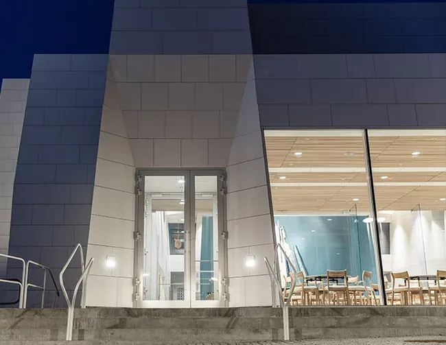 Glassdør og store vindusflater i moderne kirkebygg, med innsyn til rom med lyse trestoler.