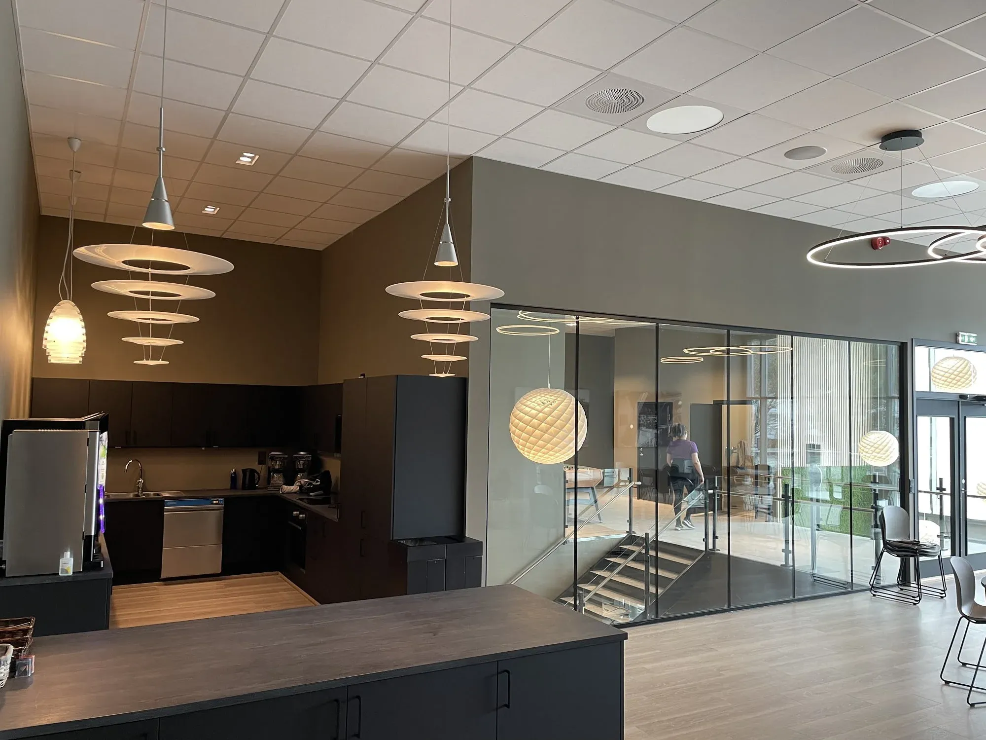 Moderne tekjøkken med mørke skap og stilige lamper, med utsikt til møterom bak glassvegg.
