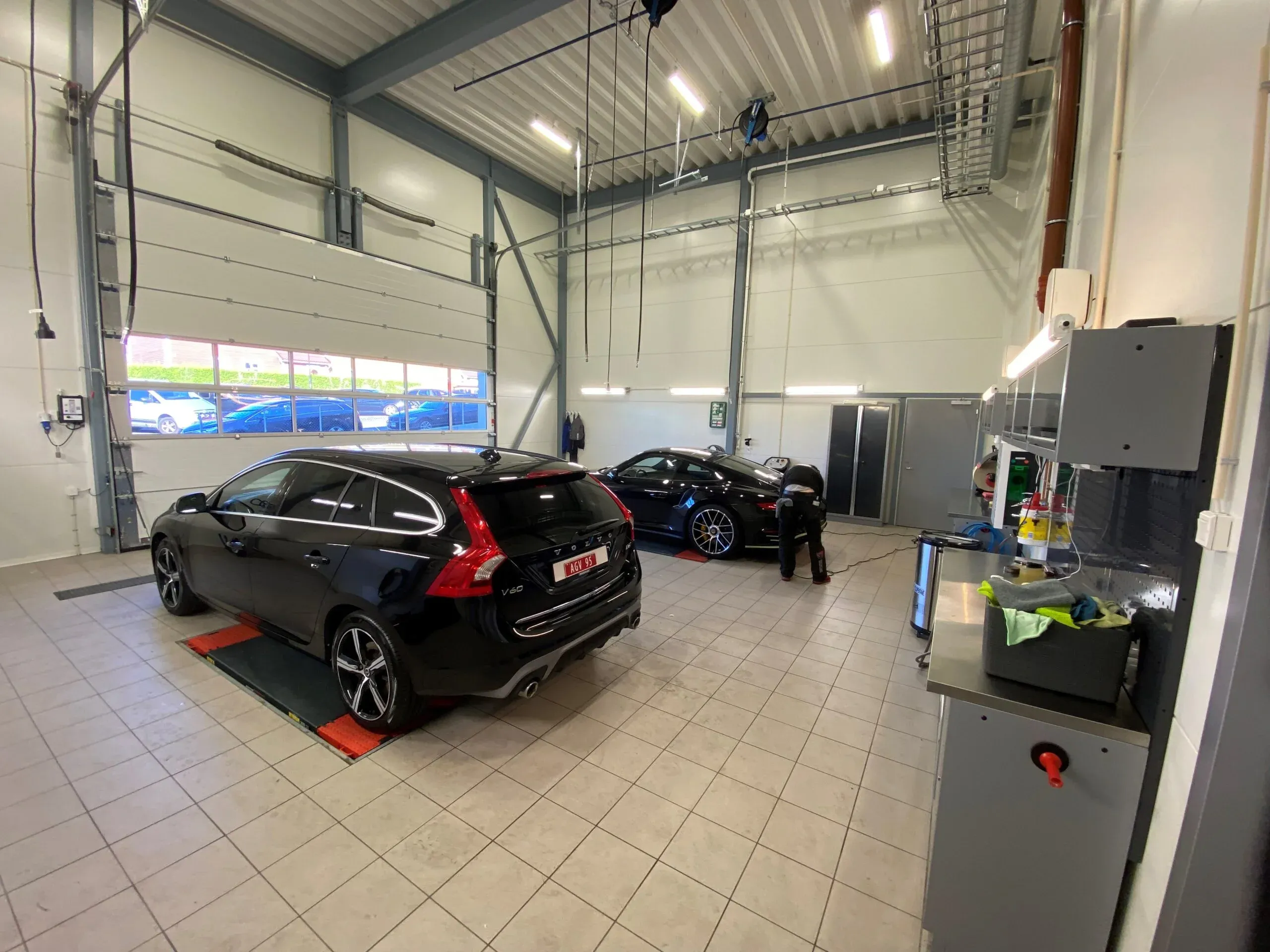 Bilpleiehall med to sorte biler og en person i arbeid, omgitt av teknisk utstyr og vaskemidler.