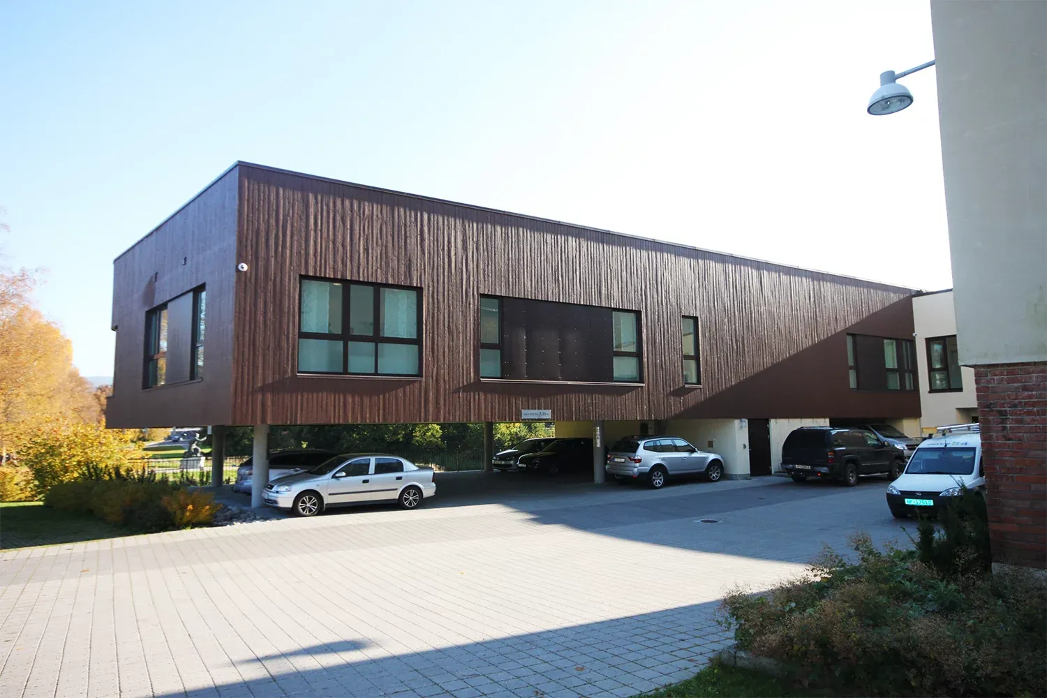 Langstrakt bygning i mørkt treverk hevet over bakkenivå, med biler parkert under og store vinduer.
