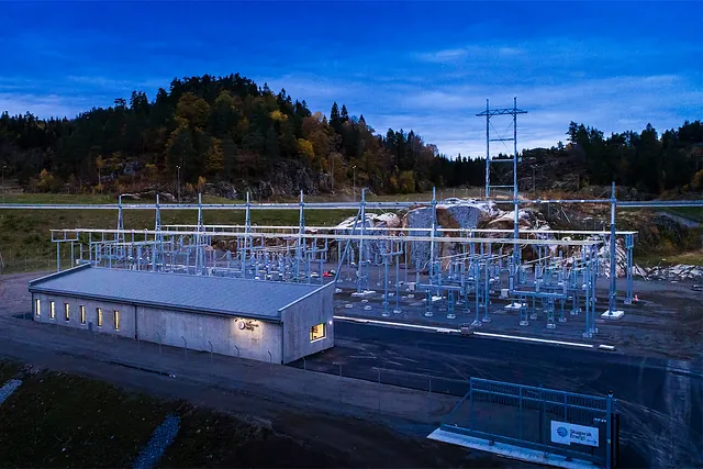 Strømstasjon i skumring med lys i bygningen, høye master og skogkledd bakgrunn.