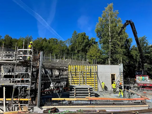 Arbeidere i refleksklær jobber med forskaling og betongarbeid ved byggeplass omgitt av skog og blå himmel.