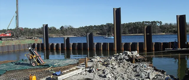 Byggeplass ved sjø med stålspunter og anleggsmaskiner, omgitt av stein og byggeutstyr.