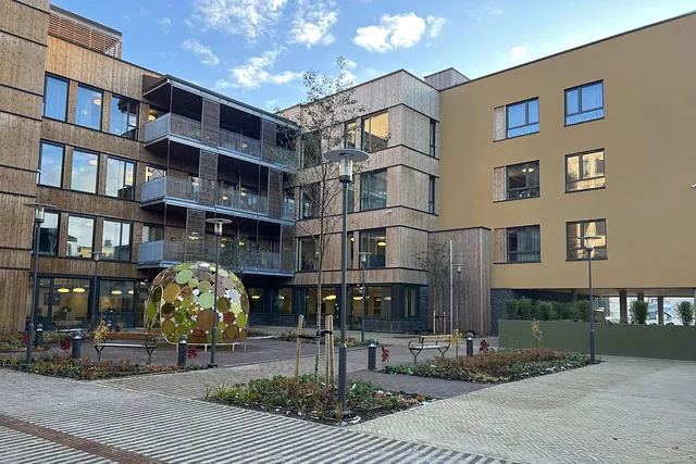 Ferdigstilt gårdsrom med benker, lamper, beplantning og kuppelformet kunstverk foran tre- og pusskledd bygg.