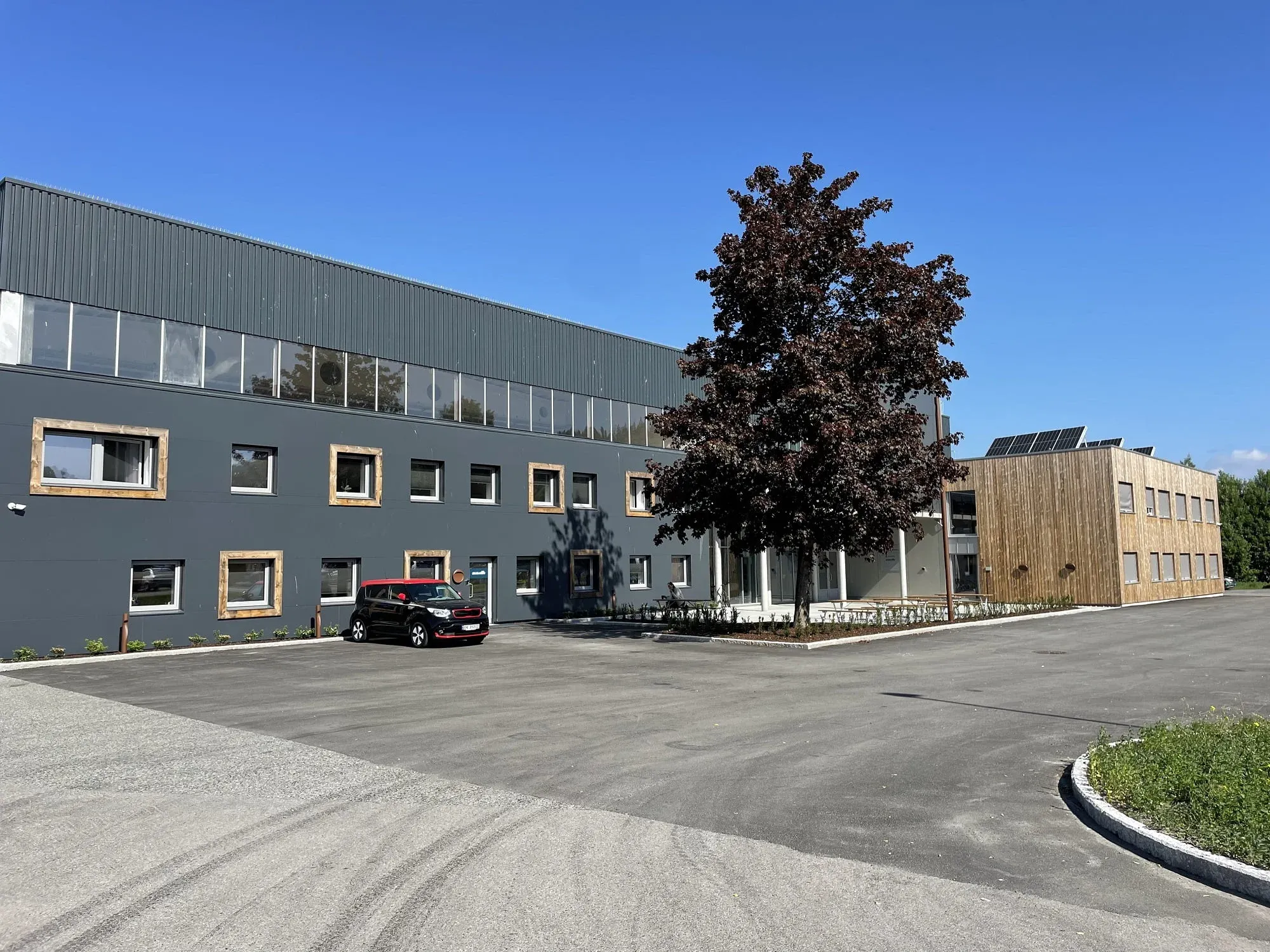 Kontorbygg i sollys med tre- og mørk fasade, parkeringsplass og trær foran inngangspartiet.
