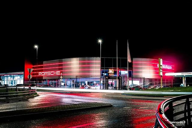 Porsche-senter belyst med røde lysspor og logo i mørket, sett fra rundkjøring med våt asfalt.