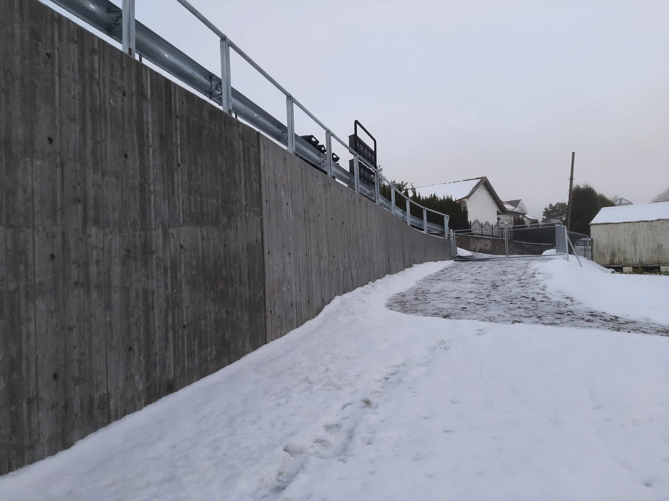 Lang, buet betongmur med snødekket gangvei som leder opp til port ved boligområde.