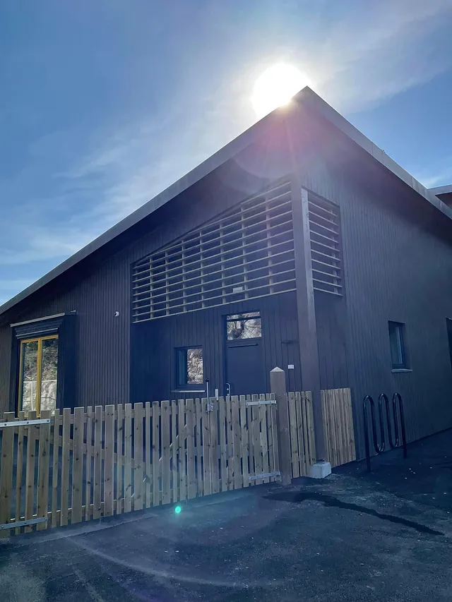 Fasade av mørkt treverk med gjerde foran og solen som skinner bak bygget.