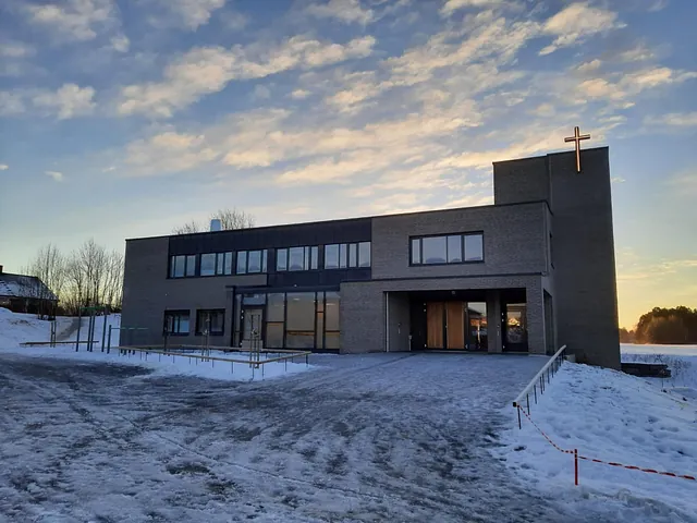 Moderne kirkebygg med lys grå teglfasade og kors på tårnet, omgitt av snø og vinterlys.
