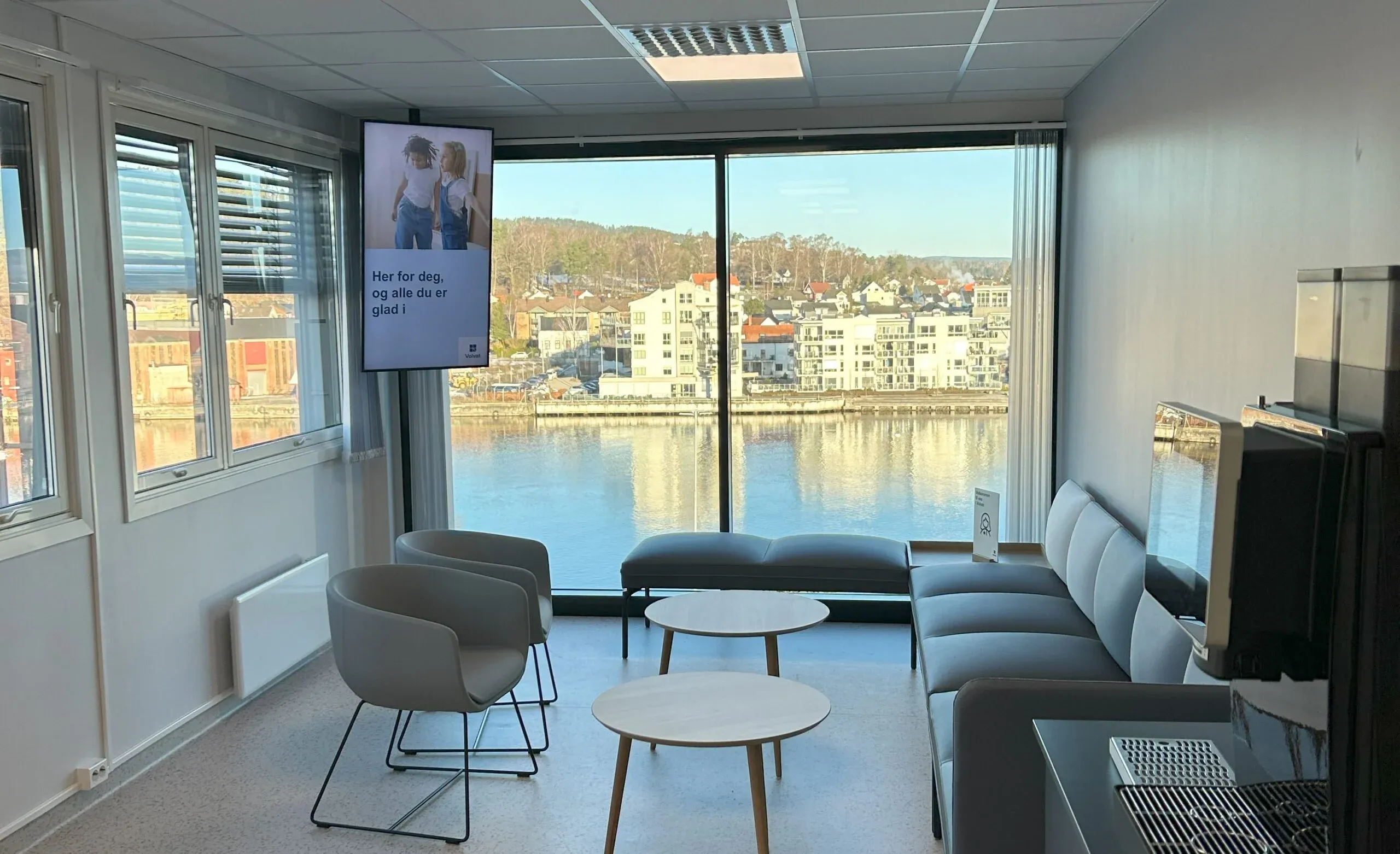 Venterom med utsikt mot elven, sofagruppe og informasjonskjerm på veggen.