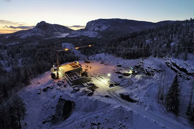 Kveldsstemning på vinterlig anleggsplass i fjellandskap, med kraner og lys som lyser opp området.
