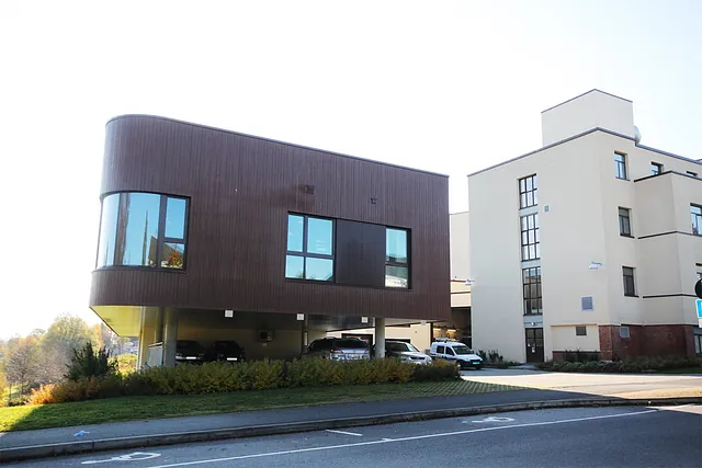 Moderne bygg med mørk trekledning på søyler, med parkeringsplasser under og store vindusflater.