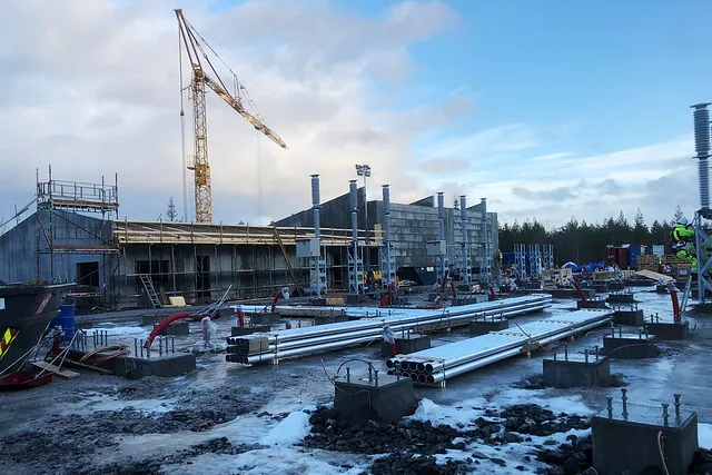Byggeplass med rørdeler, fundamenter og kran, omgitt av snø og delvis oppført betongbygg.