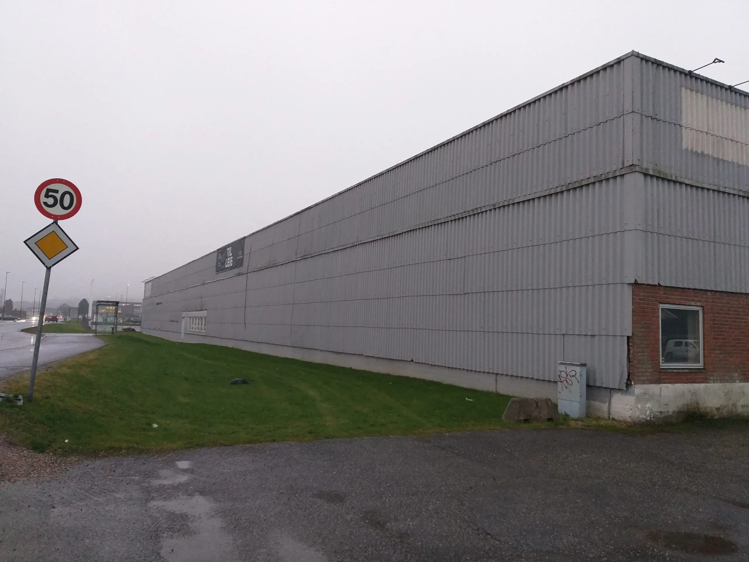 Langsiden av eldre industribygg med bølgeblikk og mur, gressplen og 50-sone skilt i forgrunnen.
