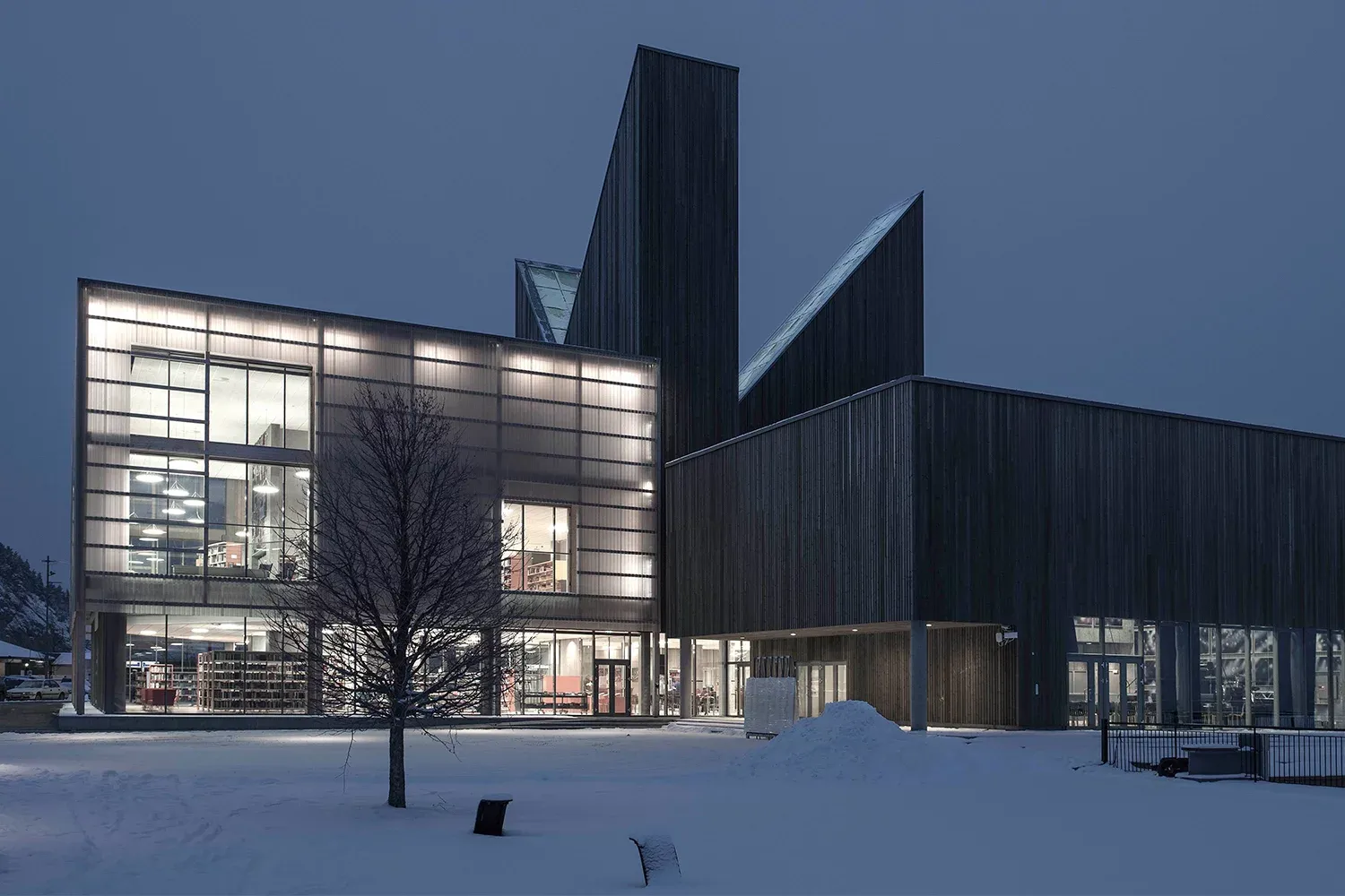 Bibliotekbygg i mørkt treverk og glass, opplyst i vinterlandskap med snø på bakken.