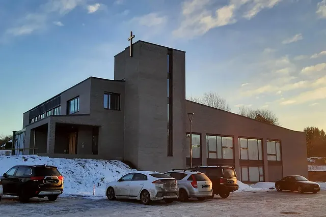 Kirken sett fra parkeringsplass i ettermiddagssol, med flere biler parkert foran bygget.