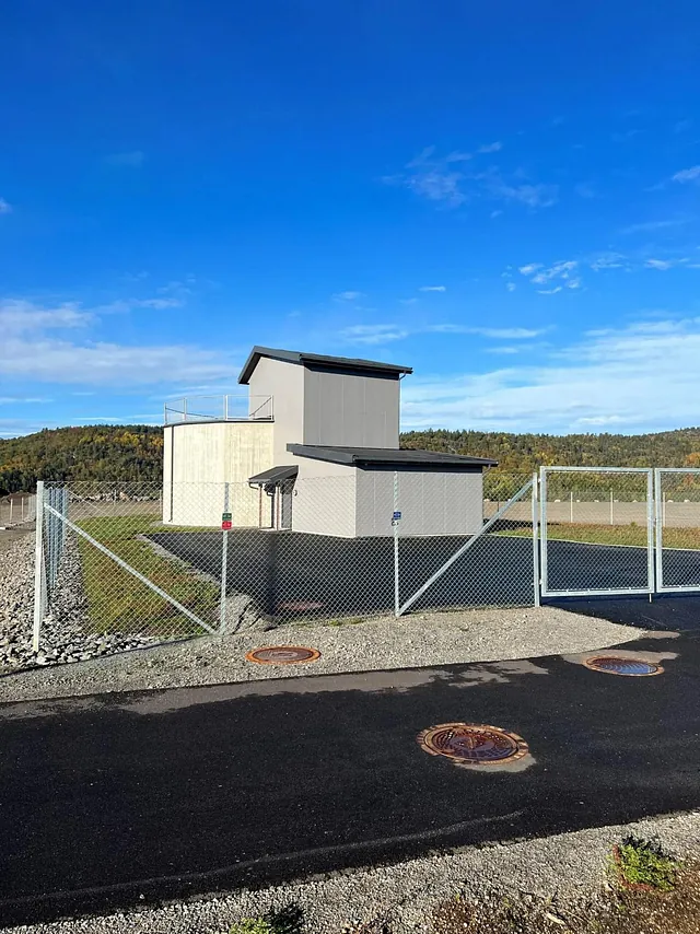 Vannbehandlingsanlegg med moderne bygg og sikret område, omgitt av natur og asfaltert vei.