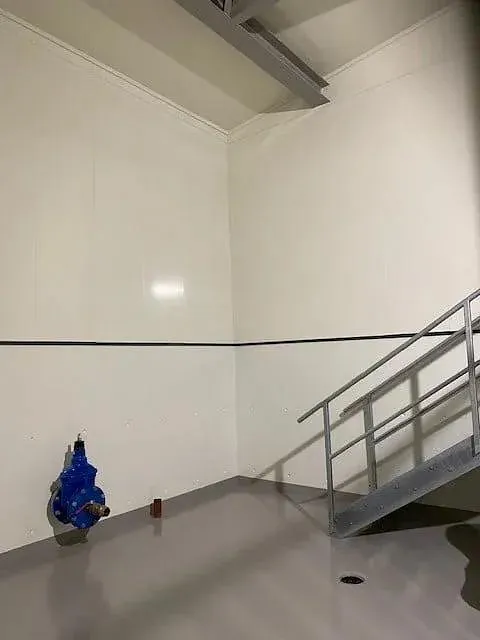 Industrielt rom med høyde, blå ventil og metalltrapp mot vegg med sluk i gulvet.