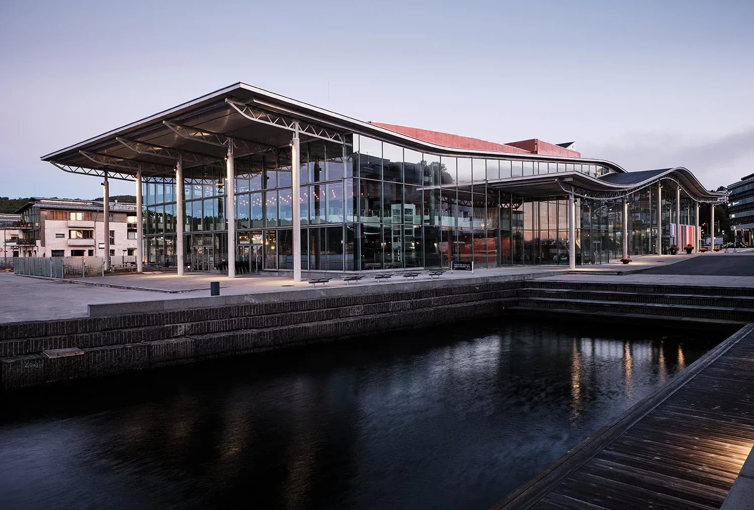 Panoramabilde av kulturhus ved vannet med stor glassfasade og bølgende tak, speilet i sjøen.
