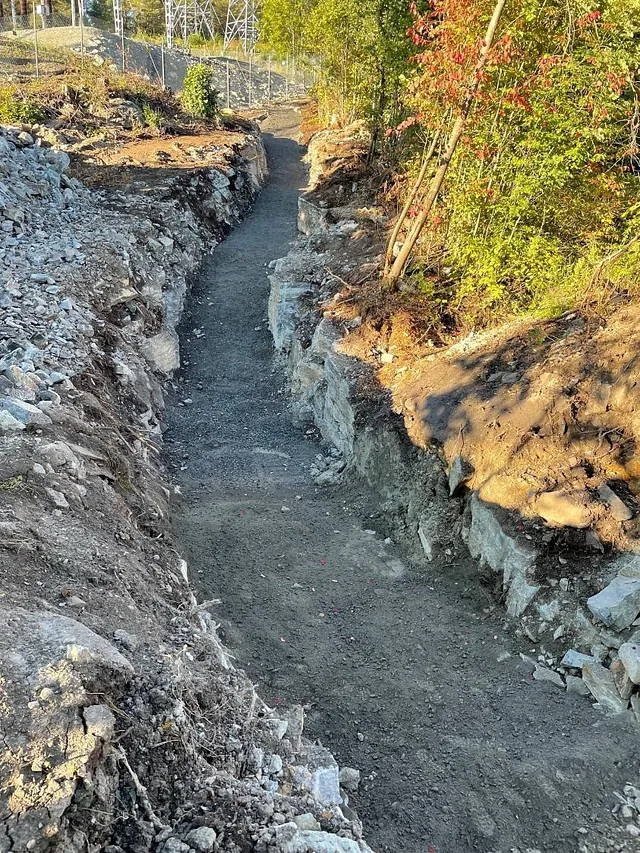 Utgravd gangsti eller trase med stein og jord, omringet av trær og fjell langs siden.