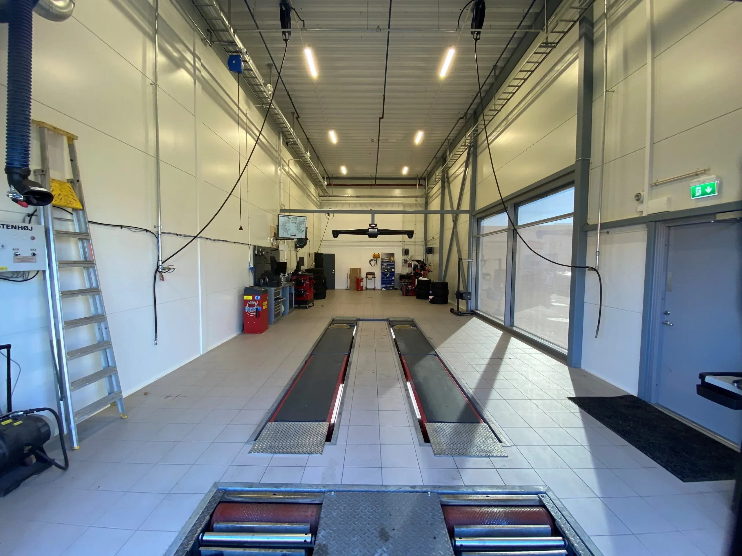 Lys verkstedhall med bremseprøverigg og utstyr for bilservice langs veggen.