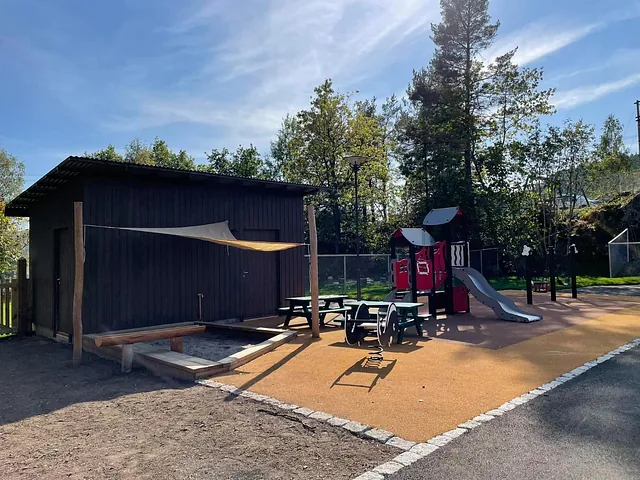 Uteområde med lekeapparater, sittebenker, sandkasse og mørk bod, omgitt av trær og gjerde.