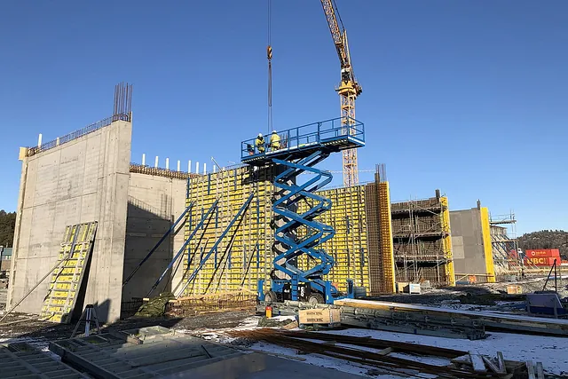 Byggeplass med heisplattform og forskalingssystem foran høy betongvegg og byggekran.