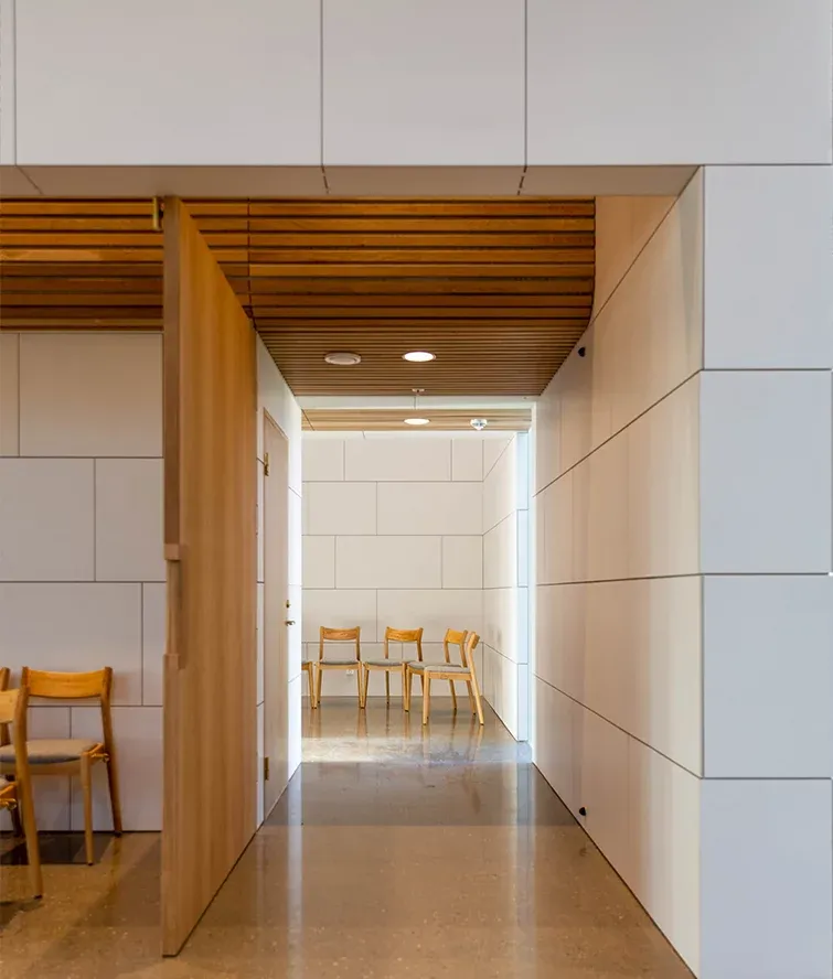 Åpen tredør leder inn til korridor med trestoler, lyse vegger og himling i trelameller.
