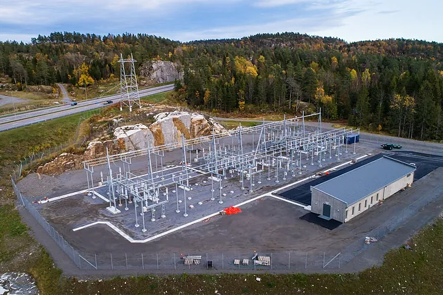 Luftfoto av strømstasjon med metallkonstruksjoner og bygning, omgitt av skog og vei i bakgrunnen.