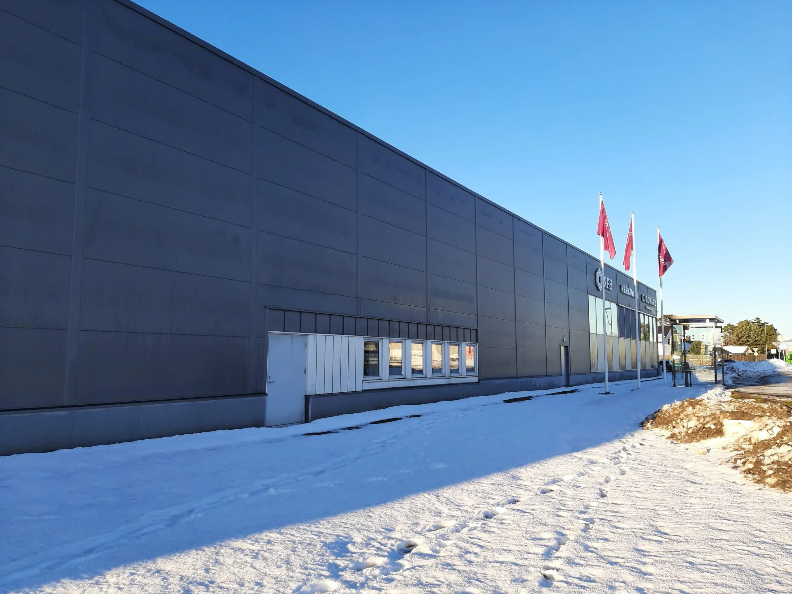 Lang fasade med mørk grå kledning og flaggstenger foran, skilt for Coward og VERKTØY.no i bakgrunnen.