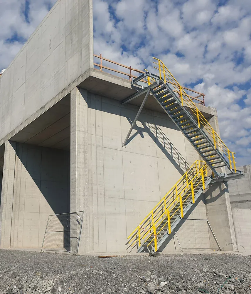 Betongkonstruksjon med utvendig trapp i metall og gule rekkverk mot en himmel med spredte skyer.