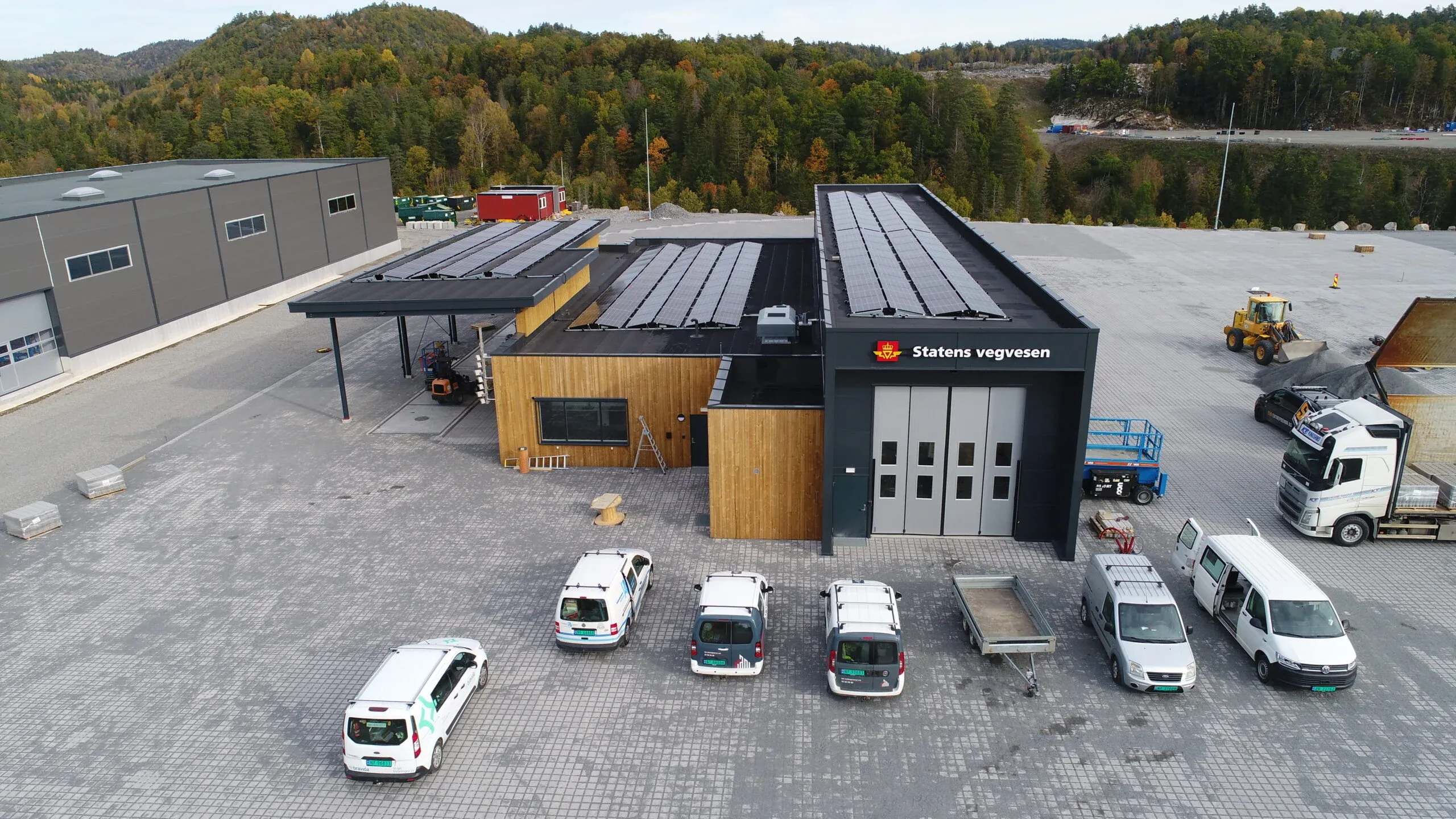 Luftfoto av Statens vegvesen-kontrollstasjon med solcellepanel på taket og flere kjøretøy parkert utenfor.