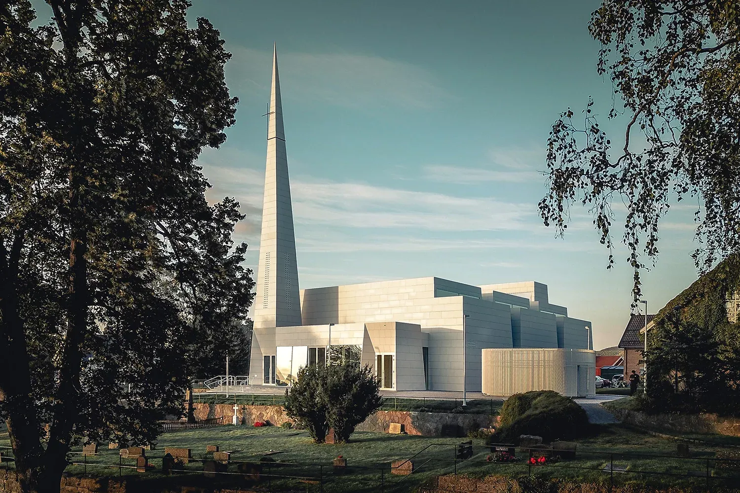 Moderne kirkebygg med høy, spiss tårnspire og lys fasade, omgitt av trær og gravlund i forgrunnen.