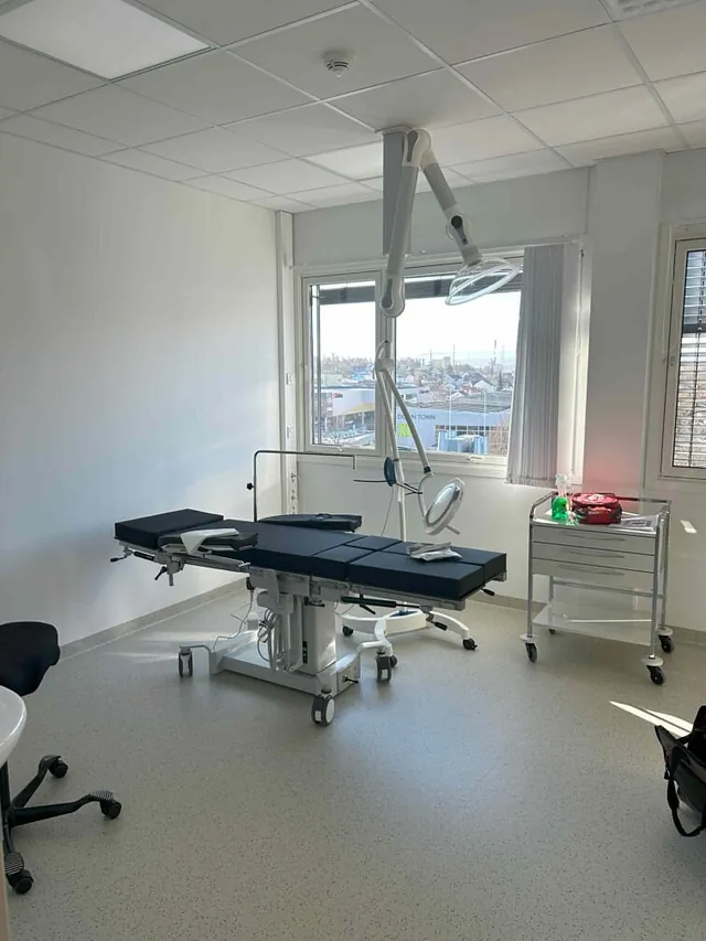 Klinisk rom med operasjonsbenk, arbeidslampe og utstyrsvogn ved vindu.