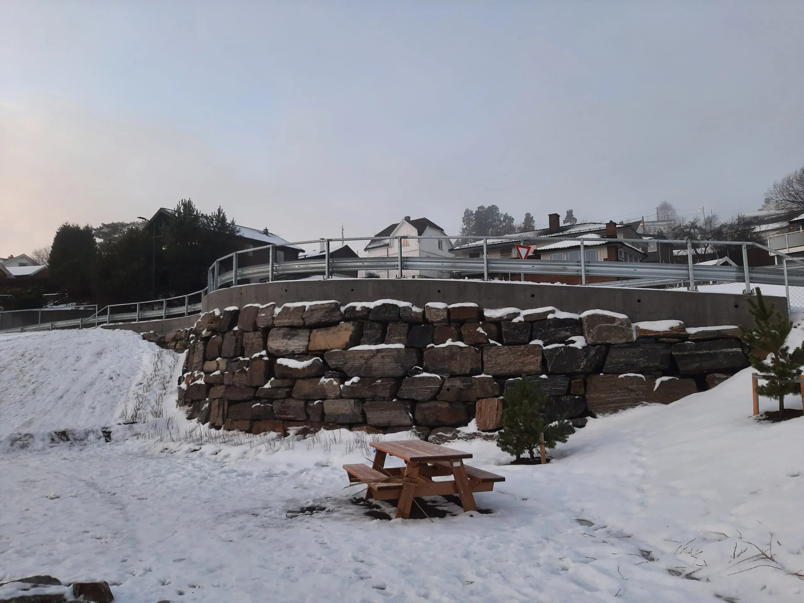 Støttemur av naturstein under betongkant og gjerde, med snødekt benk og små furutrær i forgrunnen.