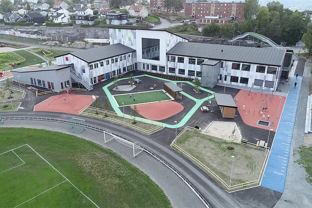 Moderne skole med lekeapparater, kunstgress og løpebane, omgitt av boligområde og grøntareal.