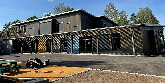 Lang fasade av barnehage med store vinduer, skygger fra pergola og lekeområde i forgrunnen.