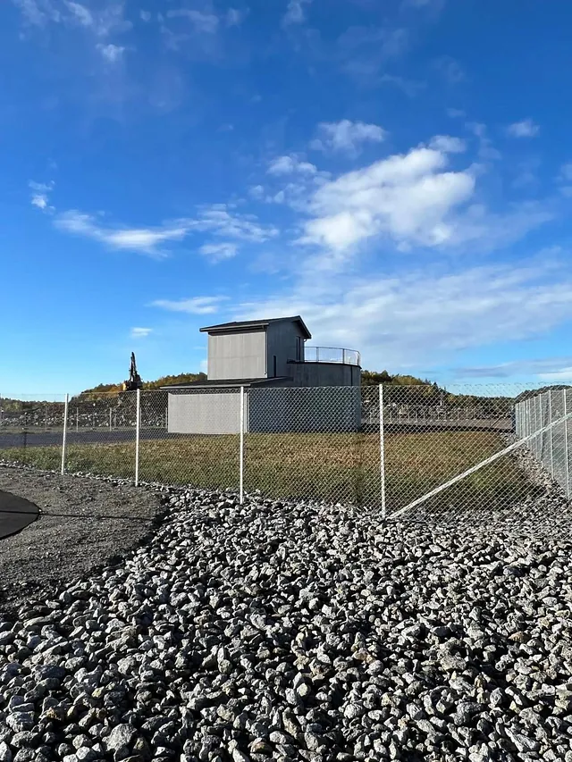 Bygning med gjerde rundt, plassert på en høyde med steinlagt grunn og blå himmel i bakgrunnen.