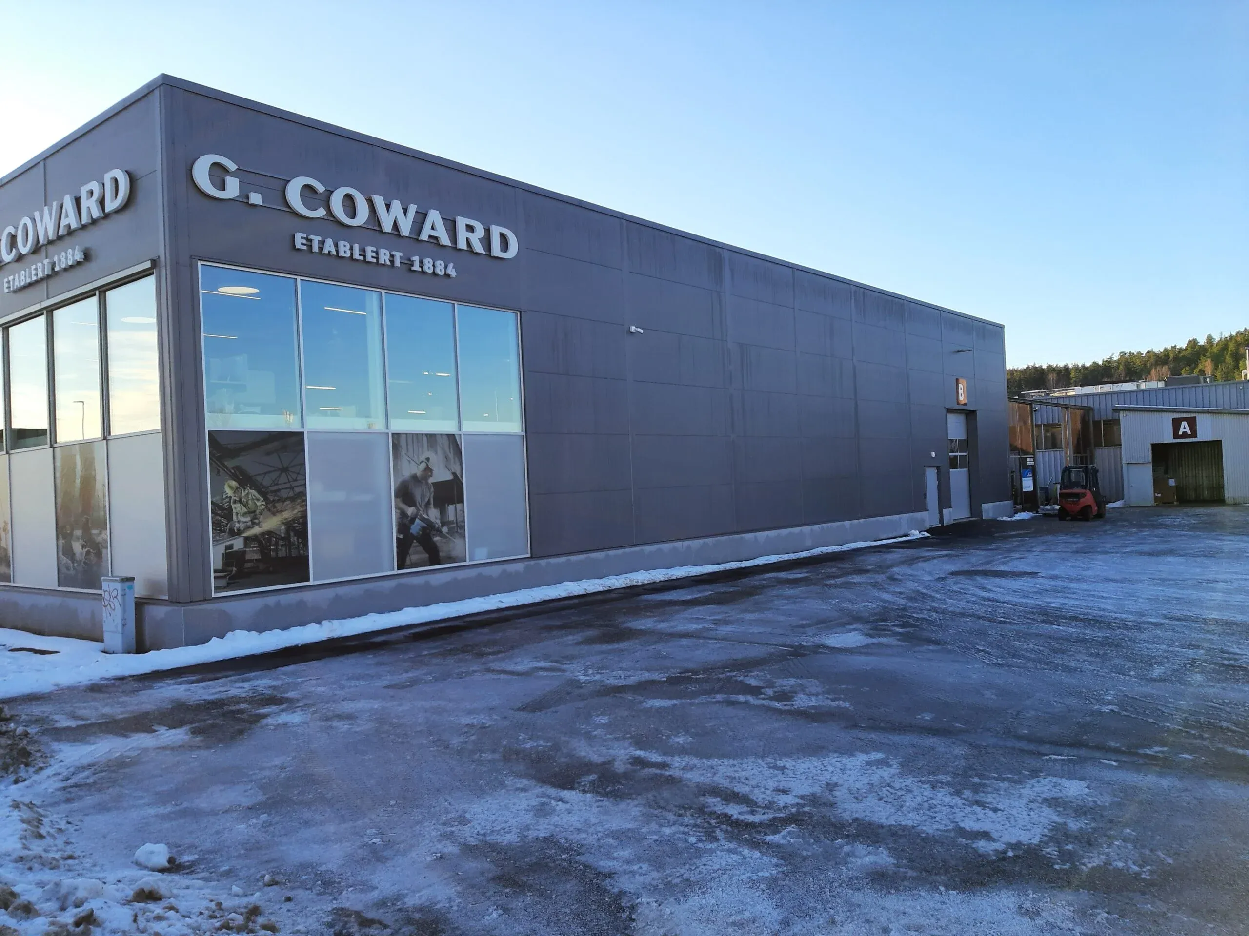 Hjørne av G. Coward-bygget med store vinduer og grå fasade, snø og is på bakken.