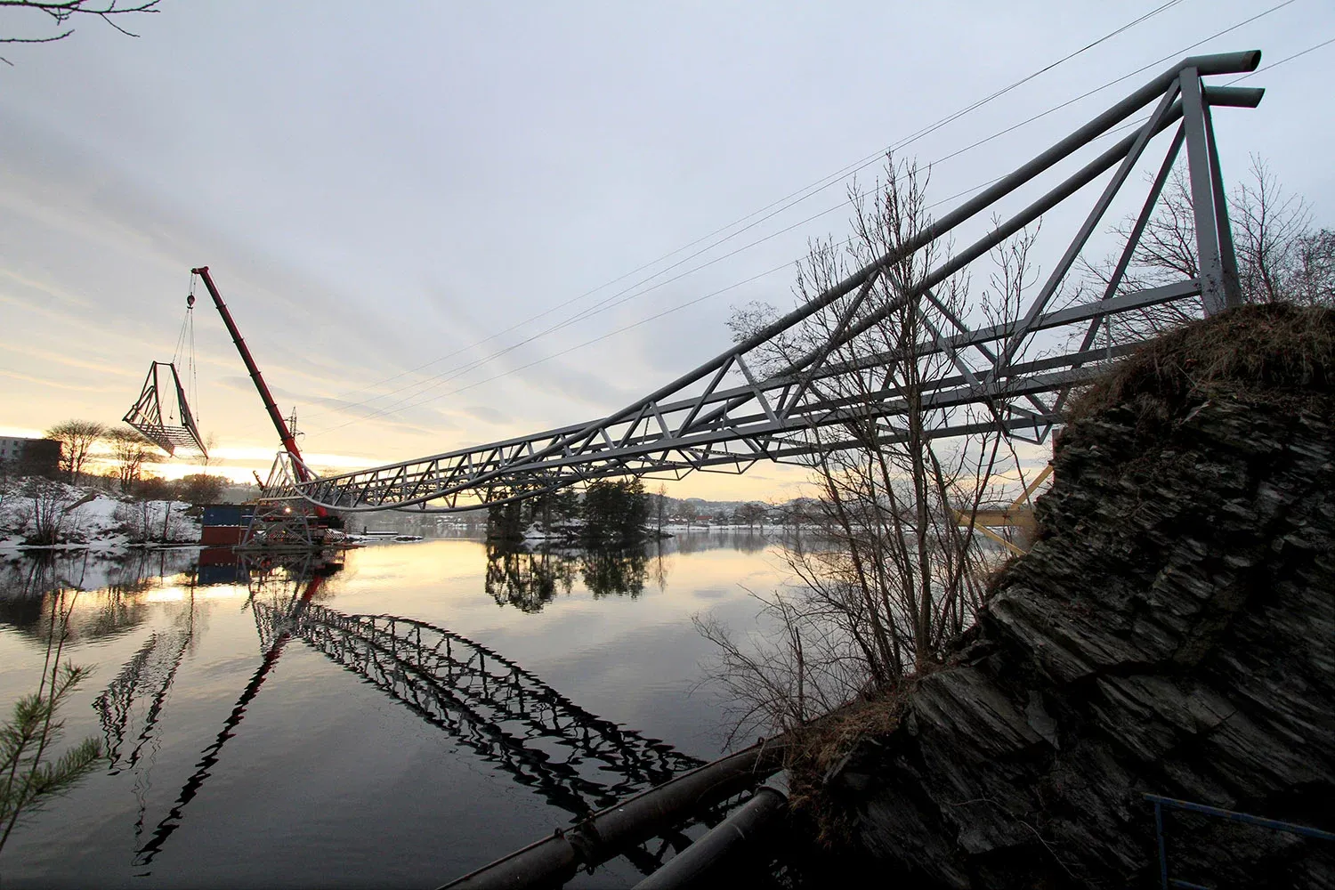 Stålbro blir heist på plass over elv ved solnedgang, med refleksjoner i vannflaten.