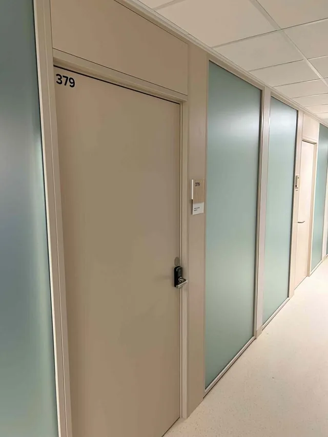 Lys korridor med dør nummer 379 og frostede glassfelt mellom behandlingsrom.