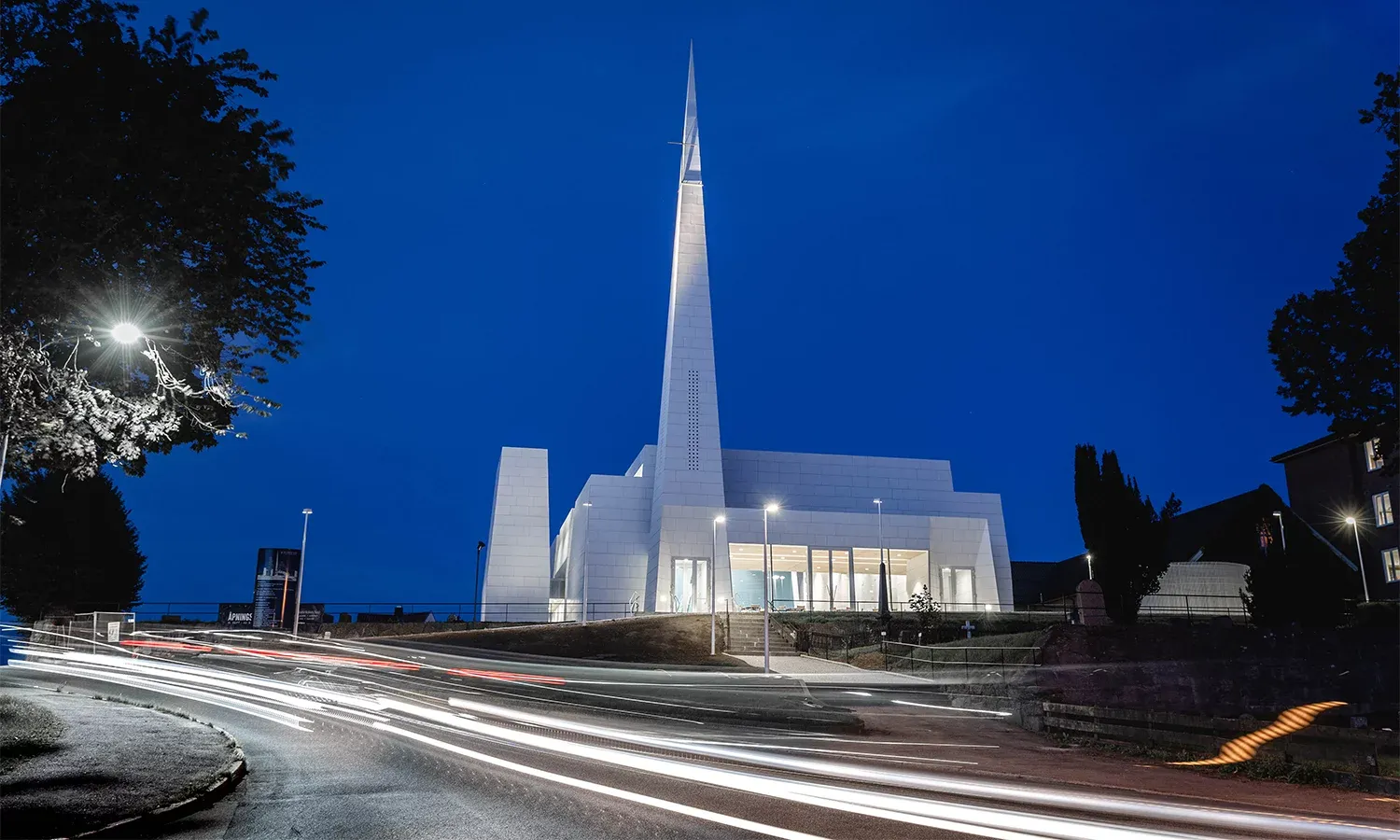 Nattbilde av moderne hvit kirke med lysende spir og lysspor fra biler i forgrunnen.
