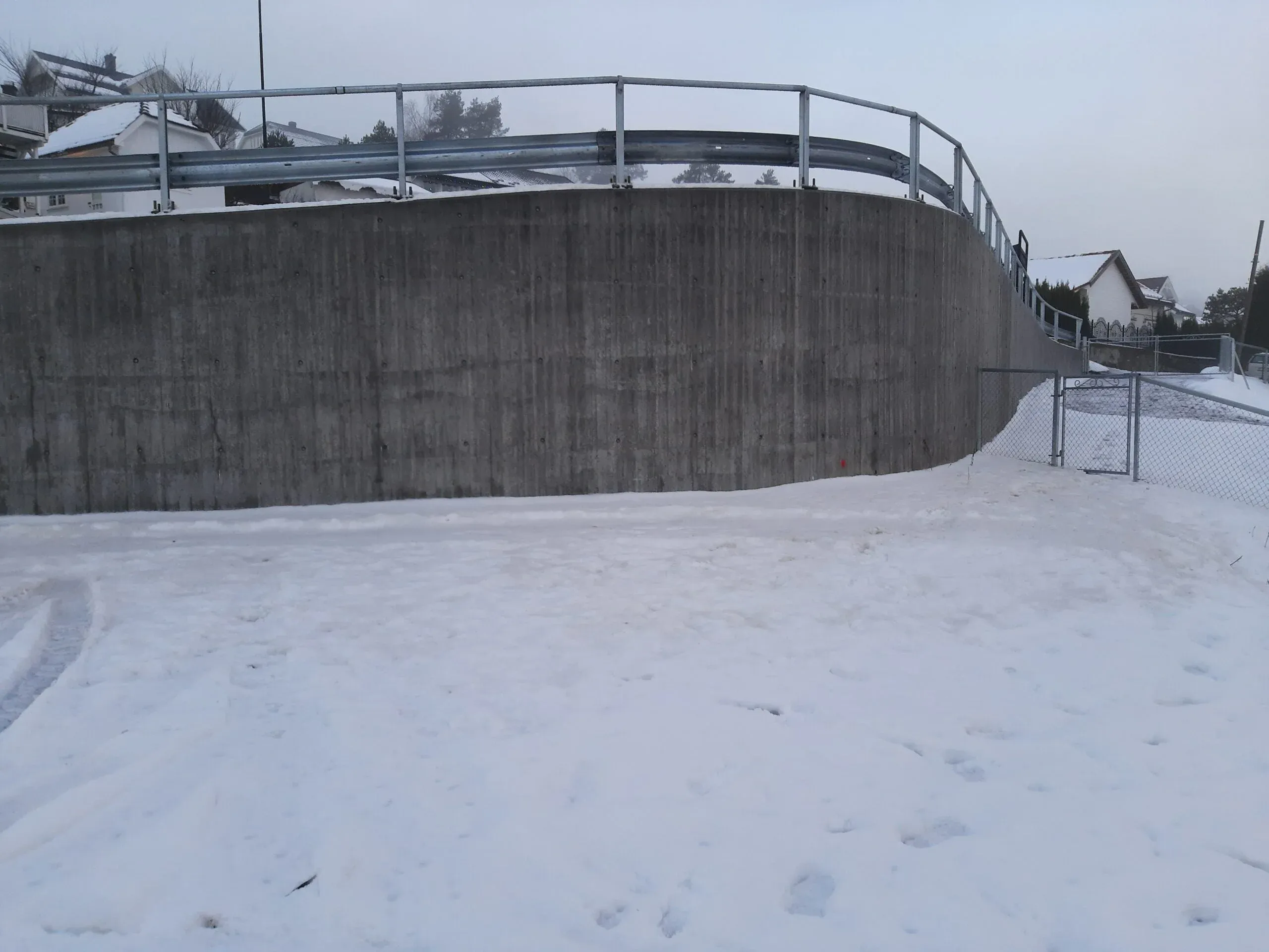 Snødekket flate foran høy, buet betongmur med autovern og gjerde, omgitt av hus og vinterlandskap.