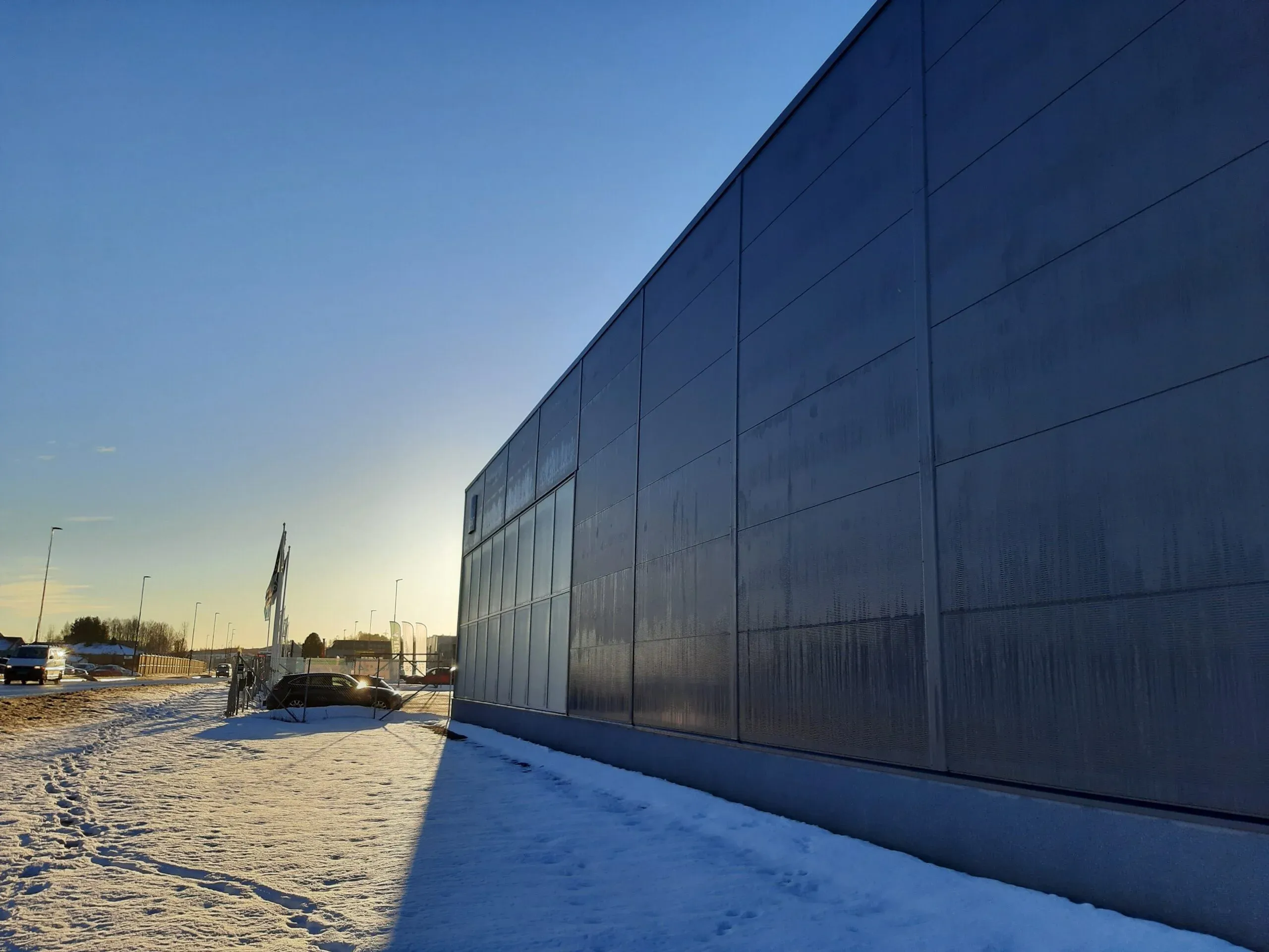 Langside av industribygg sett i motlys, med snødekket fortau og flagg ved inngjerdet område.