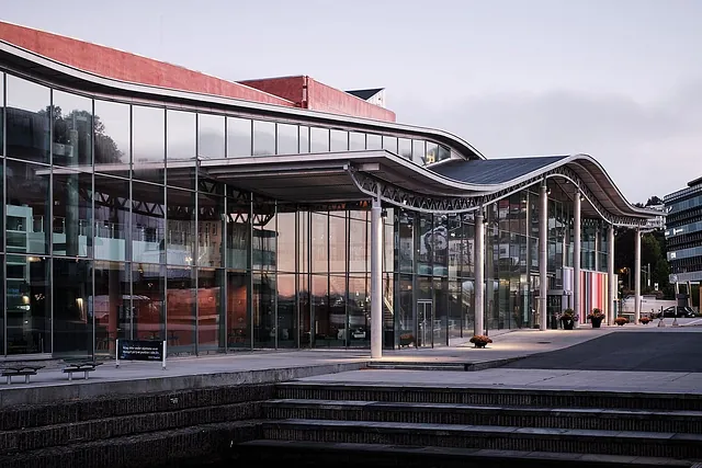 Langsiden av moderne bygg med store glassflater, søyler og fargerike detaljer mot havnepromenade.