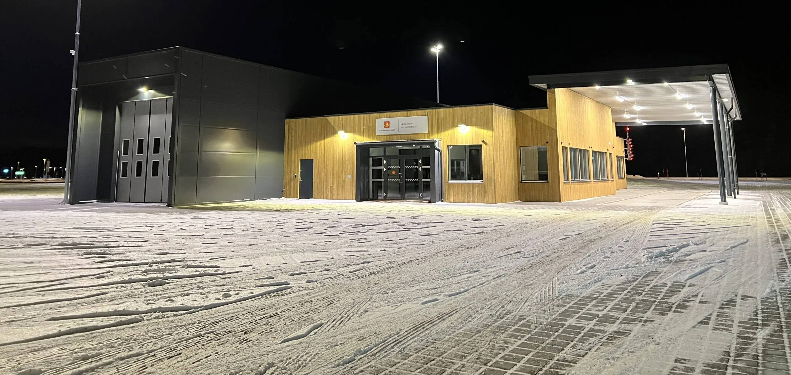 Opplyst kontrollstasjon i tre og mørk kledning om vinteren, med snødekt gårdsplass og tydelig inngangsparti.
